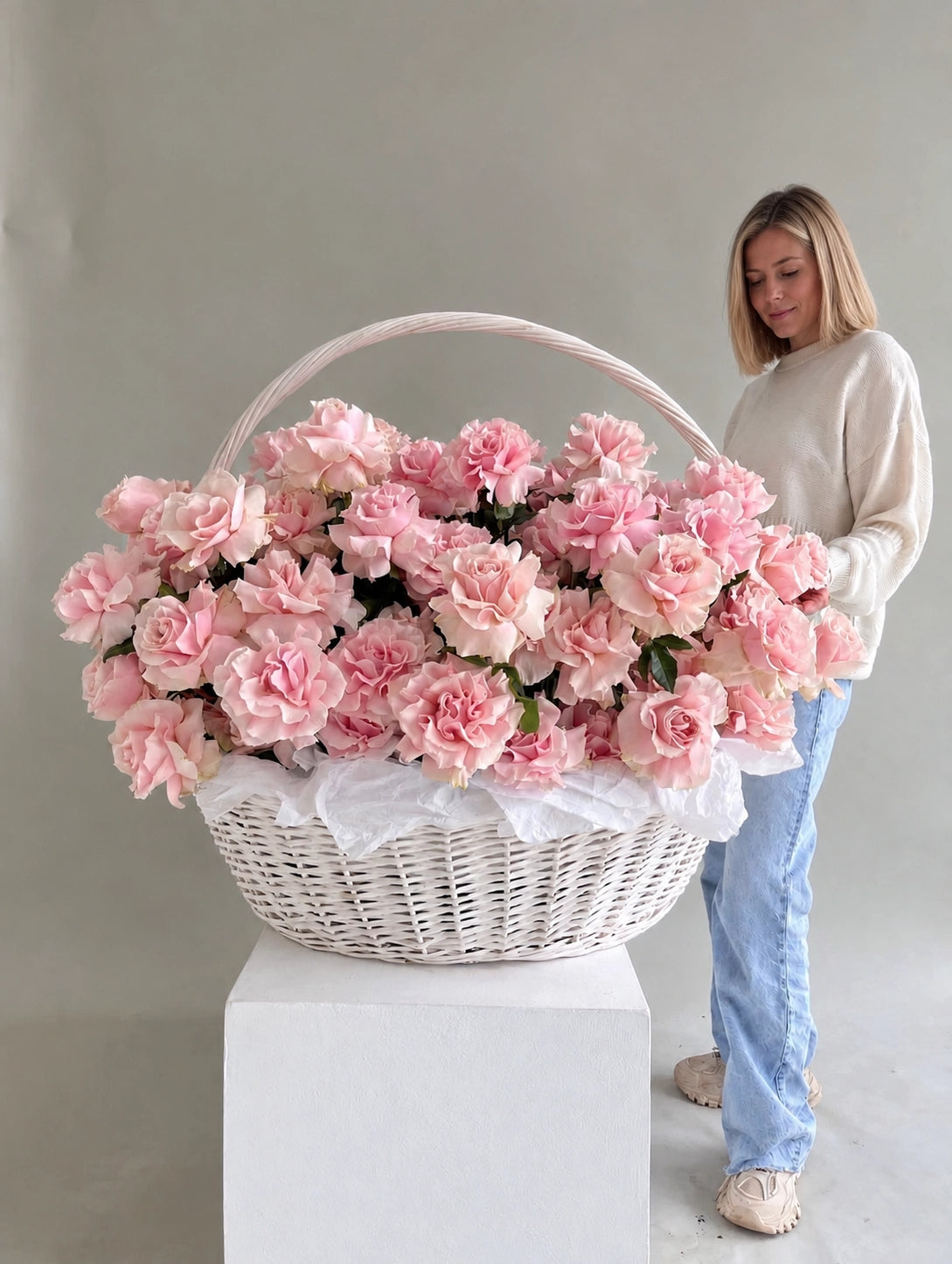 A large white wicker basket filled with pink roses, displayed on a white pedestal with a woman standing behind it against a plain light gray studio background.