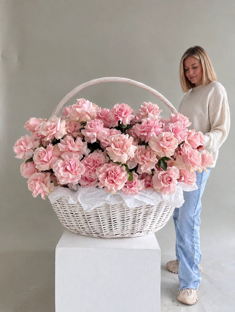 A large white wicker basket filled with pink roses, displayed on a white pedestal with a woman standing behind it against a plain light gray studio background.