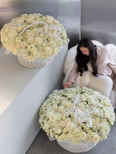 
                    
                      Two large white flower baskets filled with cream roses and white orchids are shown in a modern interior setting, with a woman seated on a white chair beside one basket.
                    
                  
