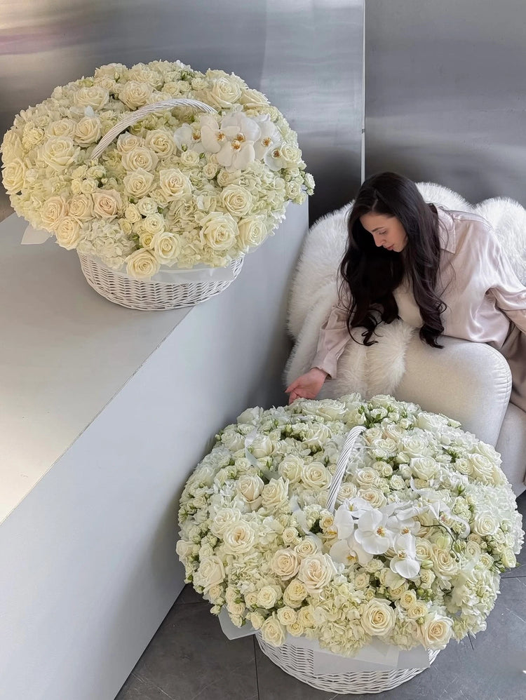 Two large white flower baskets filled with cream roses and white orchids are shown in a modern interior setting, with a woman seated on a white chair beside one basket.