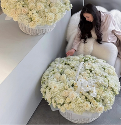 
                    
                      Two large white flower baskets filled with cream roses and white orchids are shown in a modern interior setting, with a woman seated on a white chair beside one basket.
                    
                  