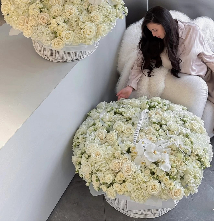 Two large white flower baskets filled with cream roses and white orchids are shown in a modern interior setting, with a woman seated on a white chair beside one basket.