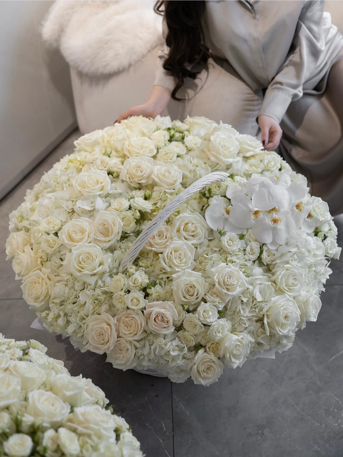 A large round floral arrangement of white and cream roses, hydrangeas, and white orchids, with a woman in a light gray satin outfit kneeling beside it on a gray floor.