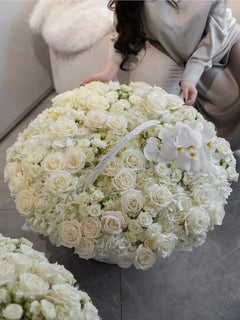 
                    
                      A large round floral arrangement of white and cream roses, hydrangeas, and white orchids, with a woman in a light gray satin outfit kneeling beside it on a gray floor.
                    
                  