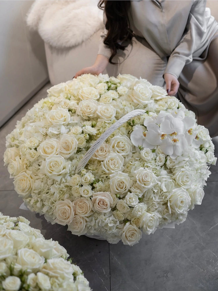A large round floral arrangement of white and cream roses, hydrangeas, and white orchids, with a woman in a light gray satin outfit kneeling beside it on a gray floor.
