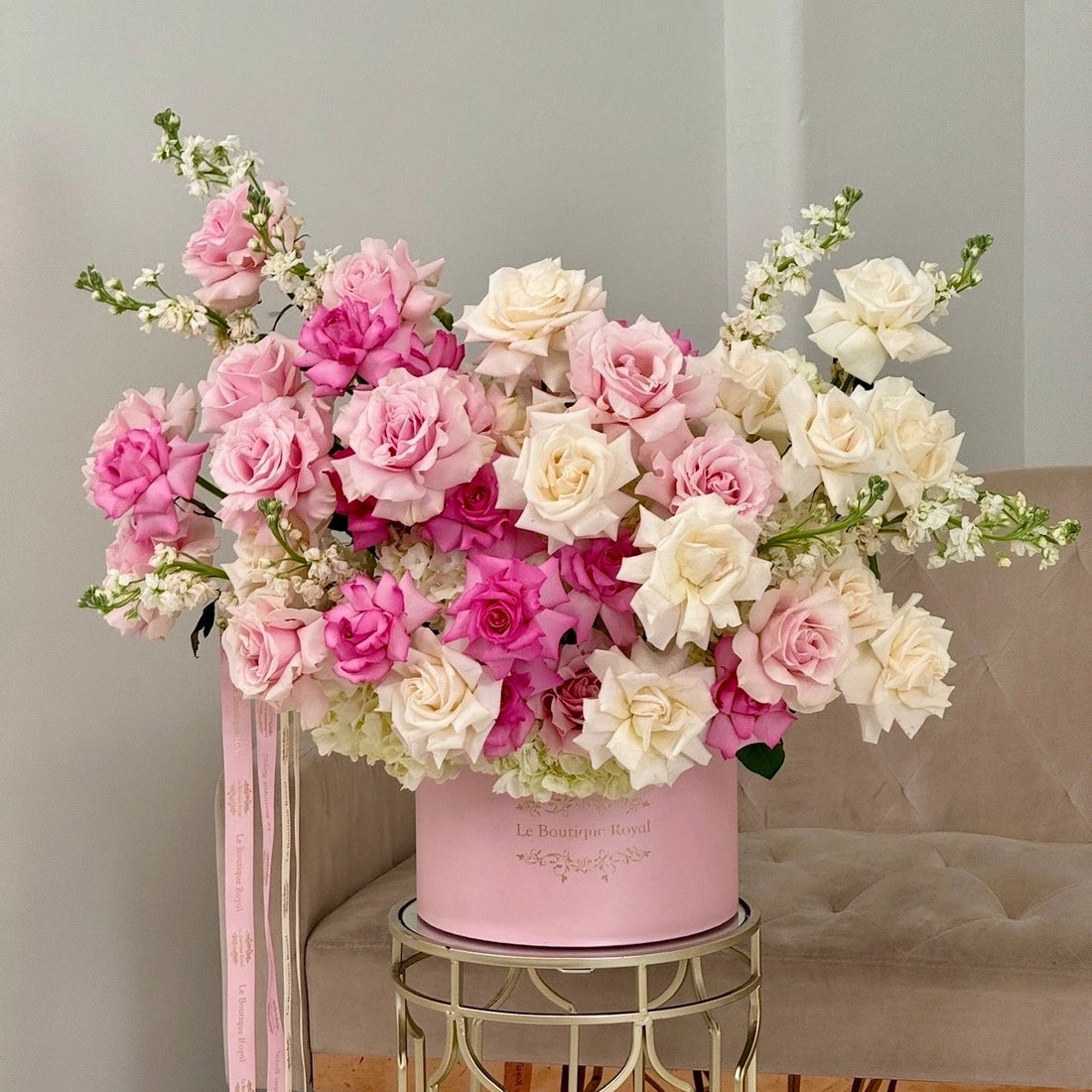 A large floral arrangement featuring pink and white roses with green stems, presented in a pink cylindrical box on a gold metal stool in front of a beige upholstered chair.
