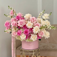 
                    
                      A large floral arrangement featuring pink and white roses with green stems, presented in a pink cylindrical box on a gold metal stool in front of a beige upholstered chair.
                    
                  